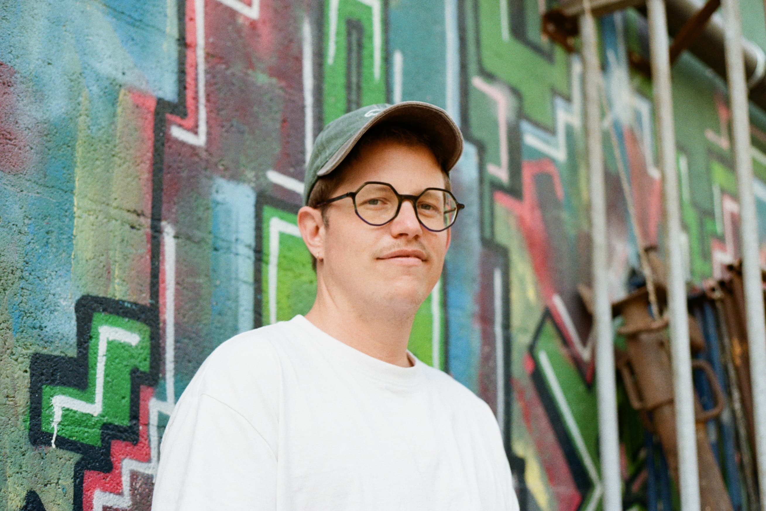 Portret van Maarten Peene, lachend in de camera voor een muur met graffiti.